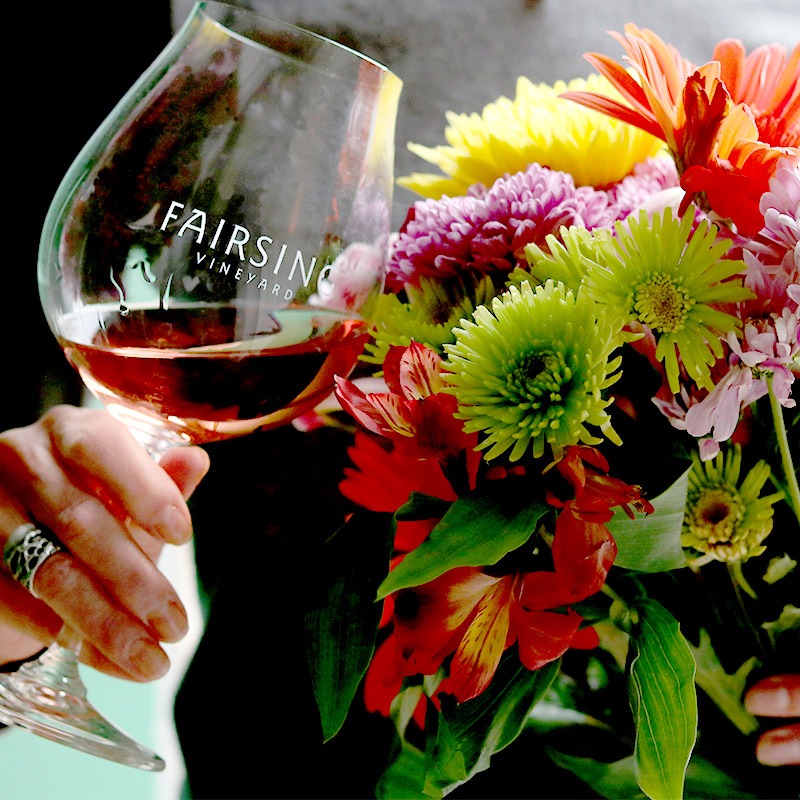 A hand holds a color bouquet of fresh flowers and Fairsing Vineyard wine glass with splash of Oregon Rosé of Pinot noir