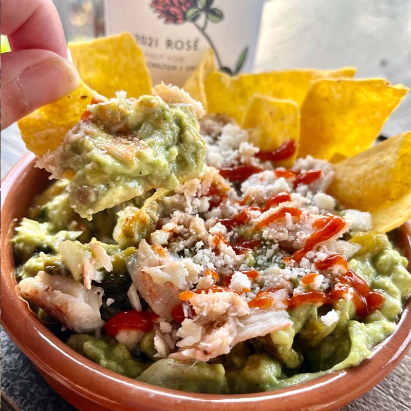 A hand dipping a tortilla chip in to Brandy Gray's Yucatan-inspired guacamole with crabmeat, queso fresco, and roasted poblano pepper with a bottle of Fairsing Vineyard Rosé of Pinot noir nearby 